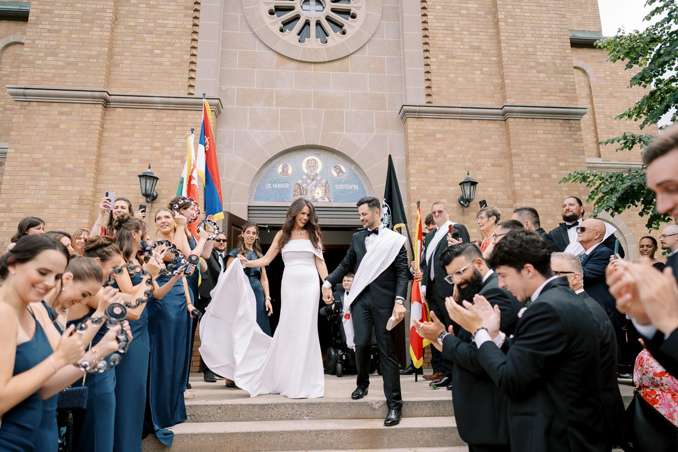 A newlywed couple joyfully exits a church surrounded by cheering guests holding tambourines, with vibrant flags in the background, capturing a festive wedding celebration.