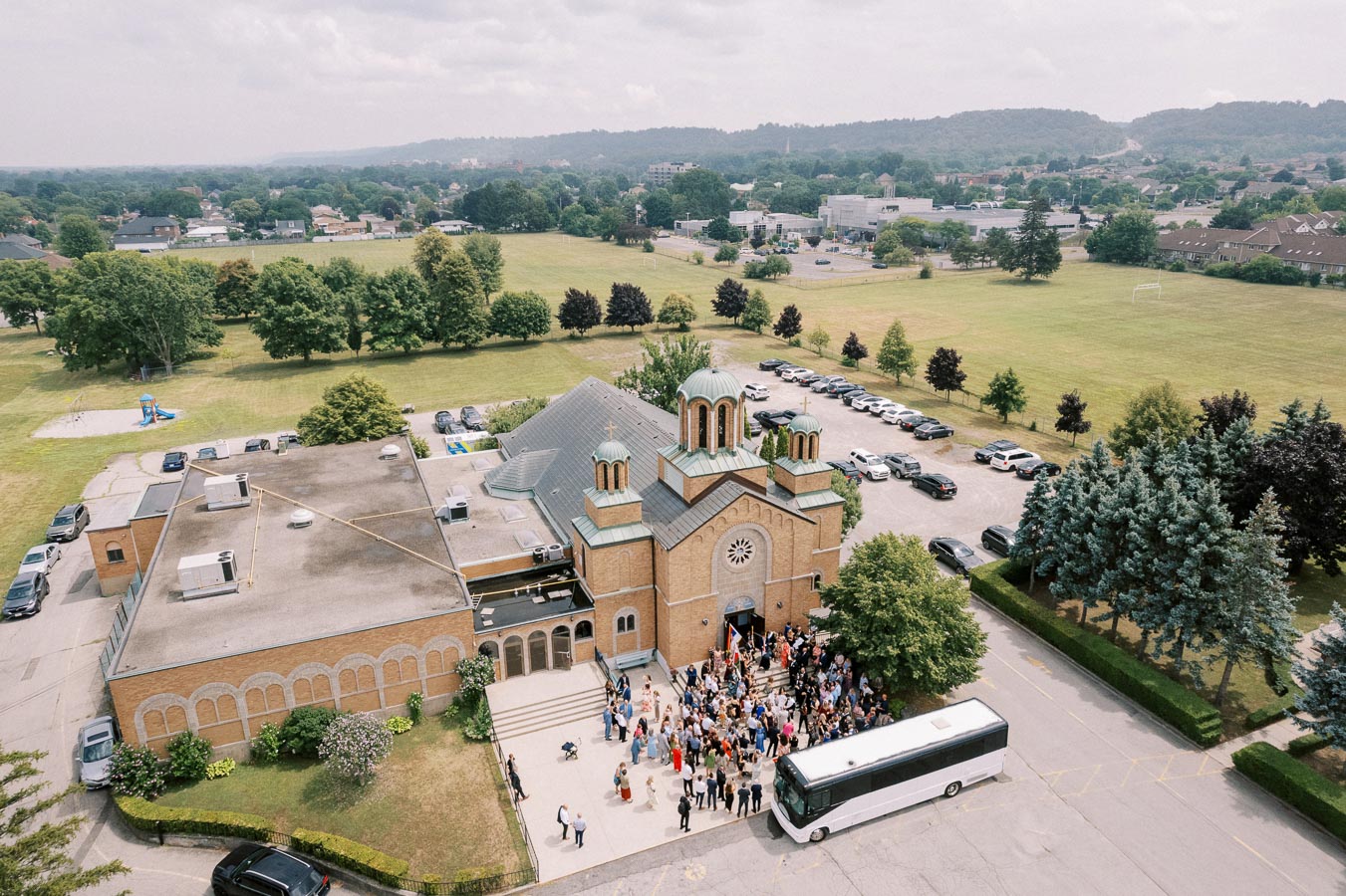 Aerial view of a large gathering outside a church with a distinctive dome architecture. A white bus is parked in front, and the surrounding area includes a spacious parking lot and lush green fields. The landscape extends to distant hills under a cloudy sky.
