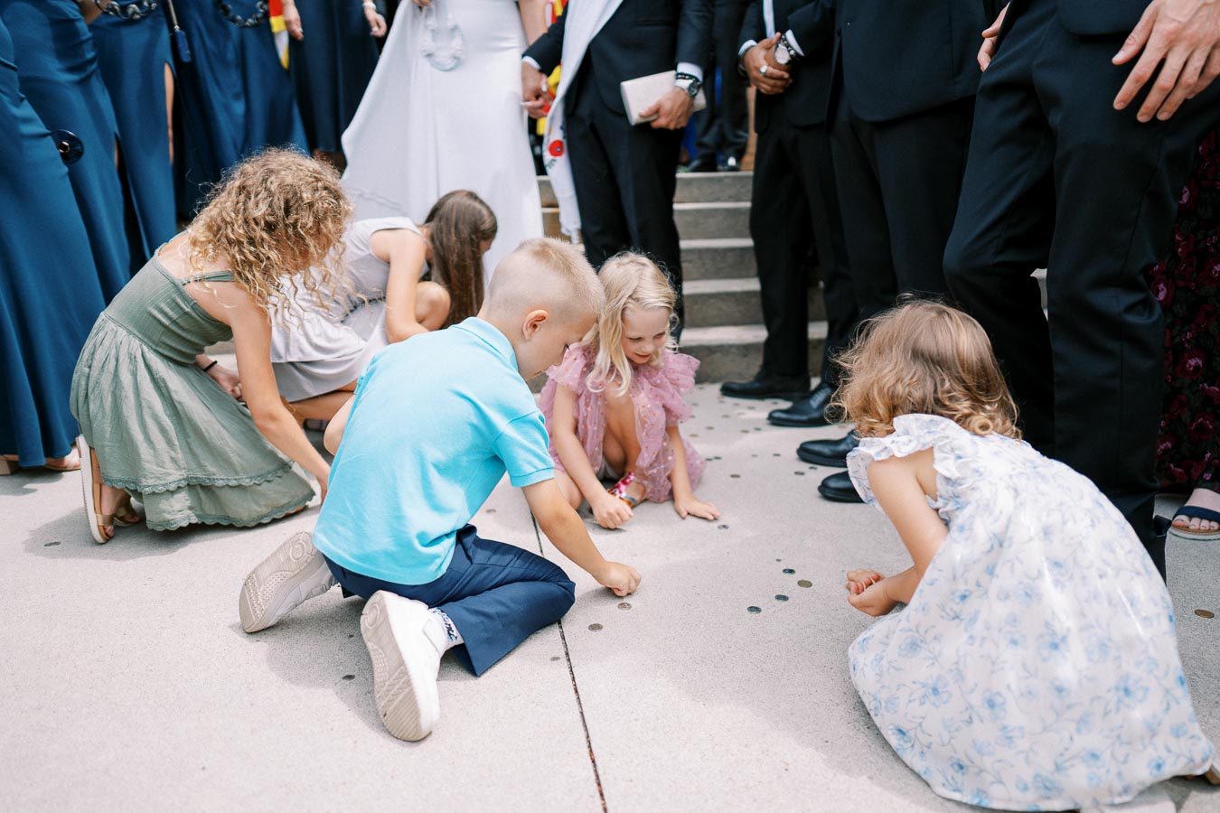 Children participating in a wedding ceremony, gathering coins from the ground, surrounded by adults in formal attire.