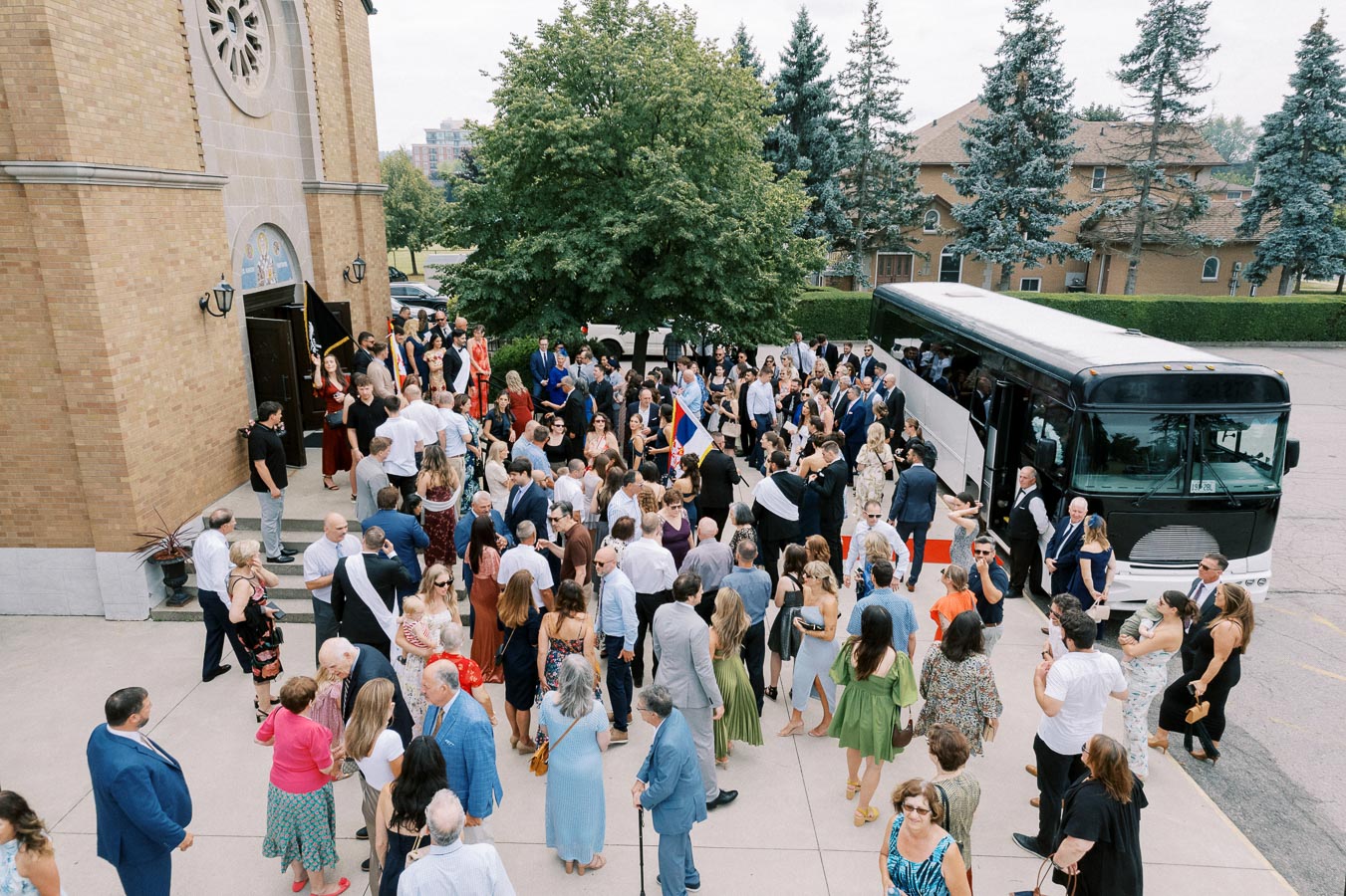 A large group of people gather outside a church, dressed in formal attire, next to a parked bus, indicating a wedding or special event in a vibrant social setting with greenery in the background.