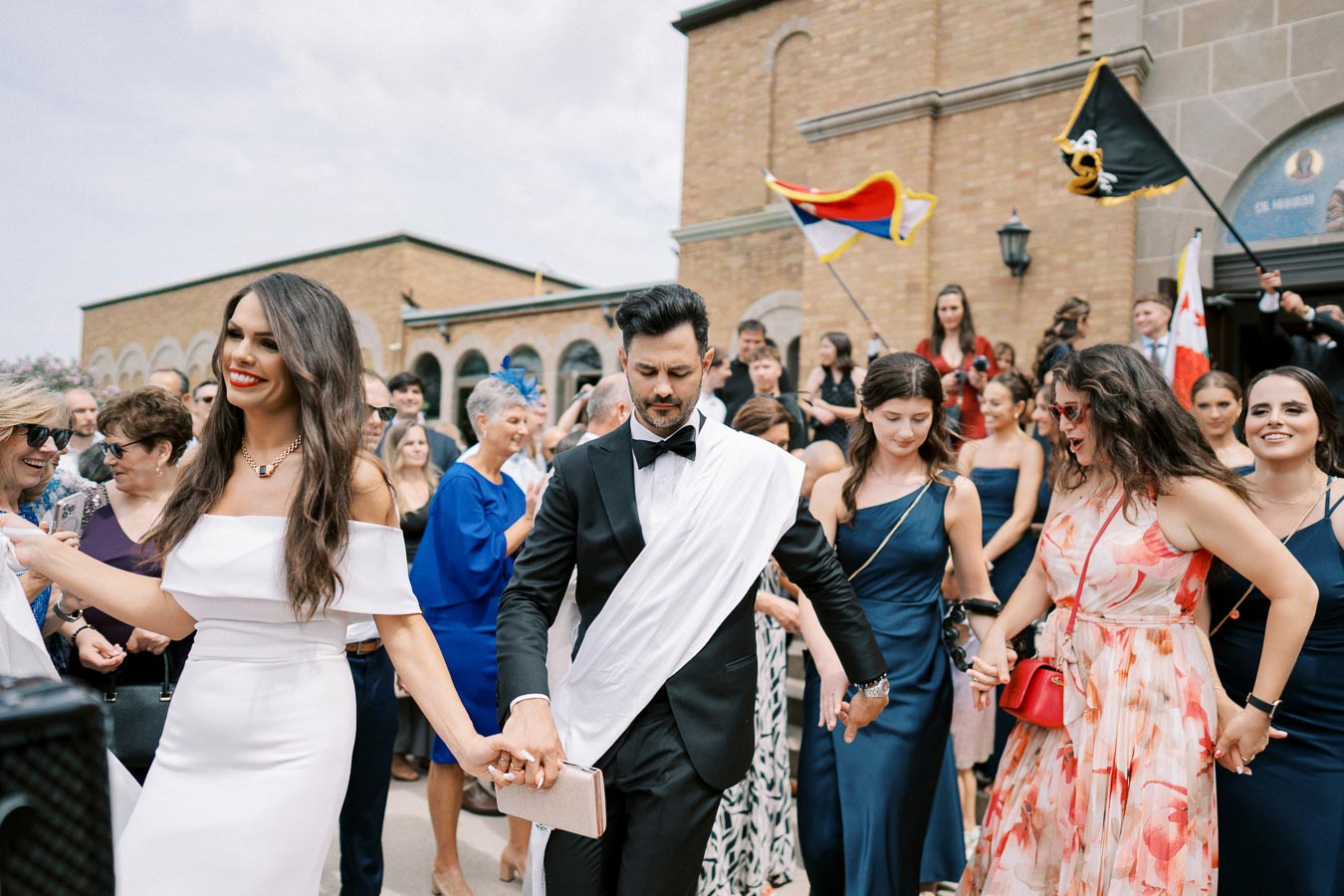 A wedding celebration with a couple in formal attire surrounded by joyful friends and family, holding hands and dancing outside a historic building. Guests are dressed in elegant outfits, and flags are being waved in the background.