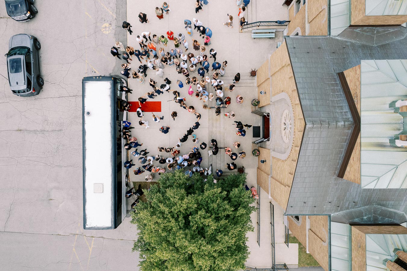 Aerial view of a large crowd gathering outside a building with a red carpet leading to a bus, surrounded by parked cars, showcasing a bustling event or gathering.