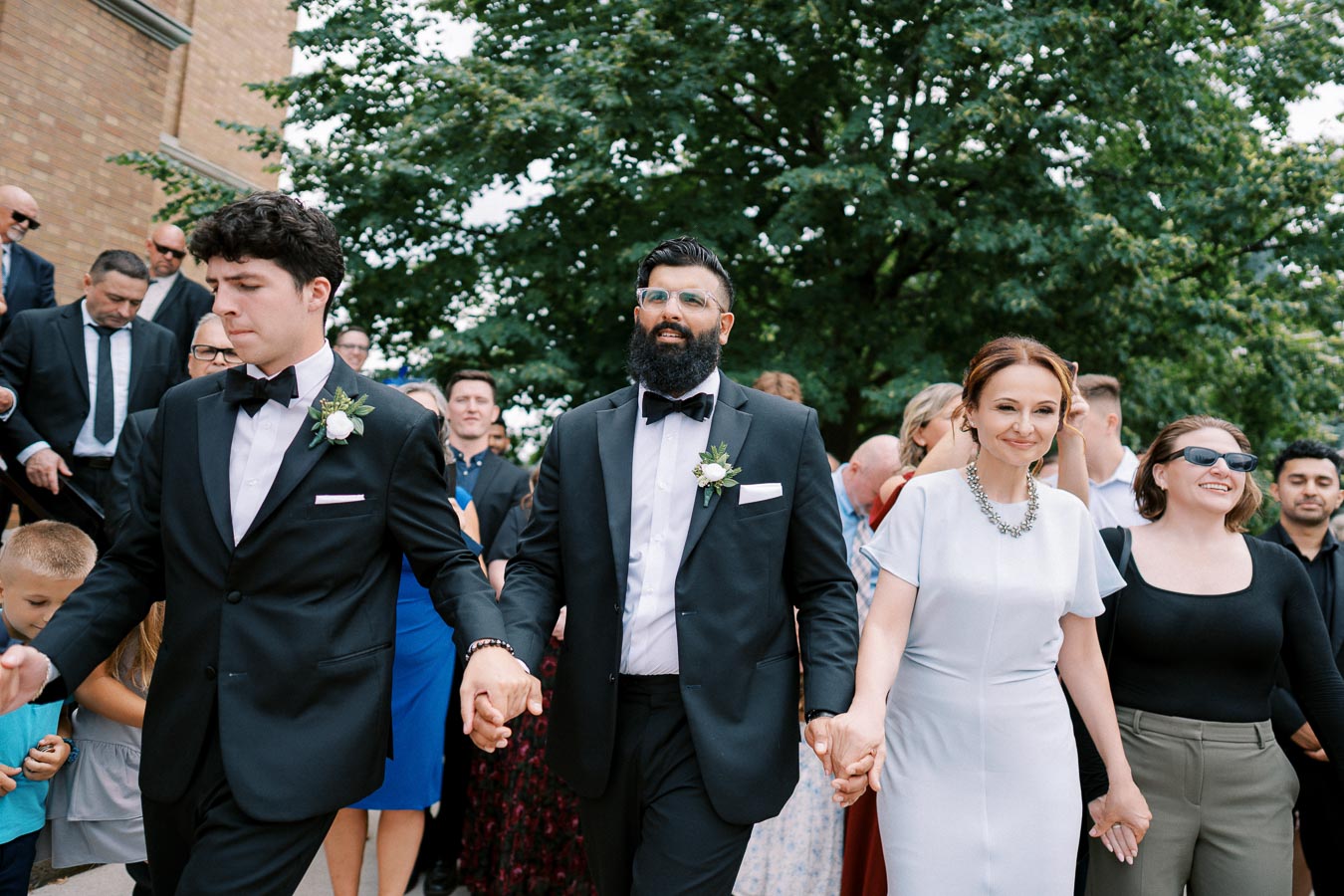 A group of people in formal attire attending a wedding celebration, with two men in black suits and a woman in a white dress, surrounded by happy guests outdoors.