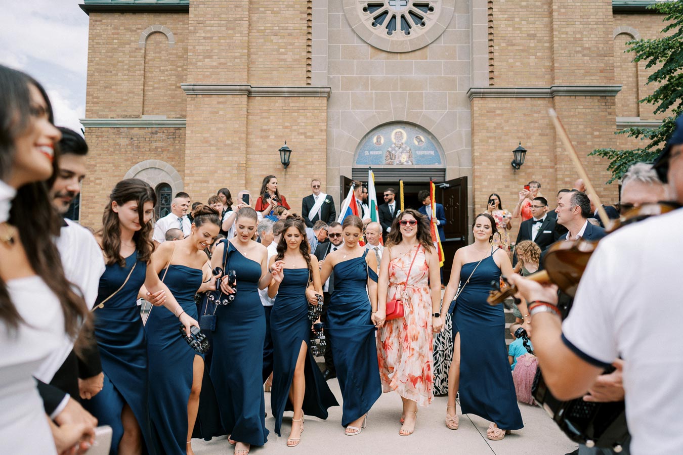 Group of people in formal attire, including women in blue dresses and a woman in a floral dress, gathered outside a church with a violinist playing.