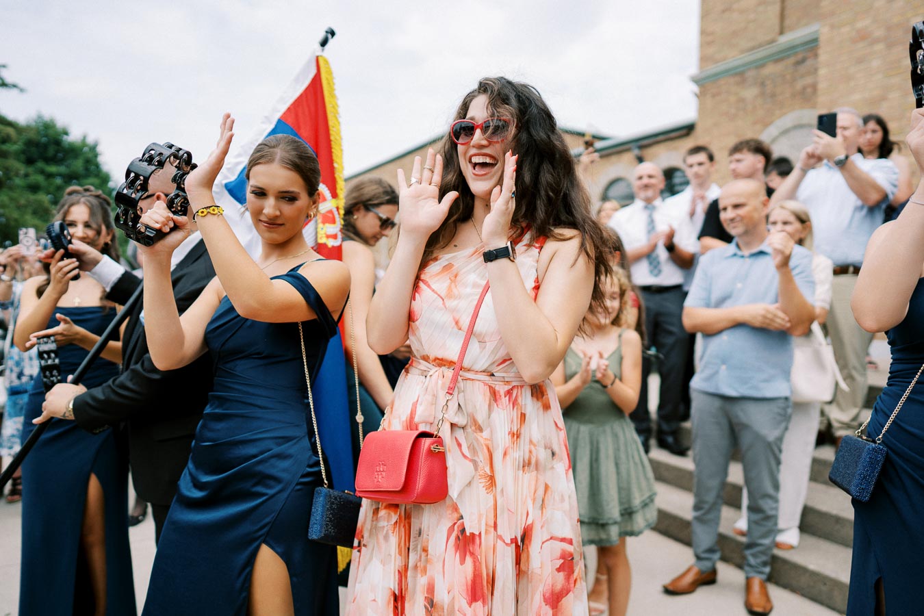 Group of people celebrating outdoors with a tambourine and flag, featuring women in elegant dresses and a joyful atmosphere.