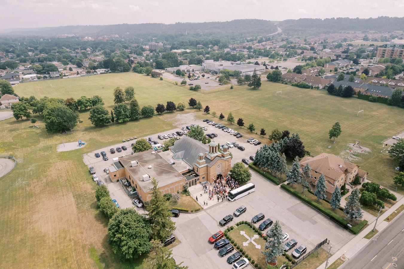 Aerial view of a large gathering outside a church surrounded by a spacious parking lot and lush green fields, showcasing a serene suburban neighborhood with distant rolling hills in the background.