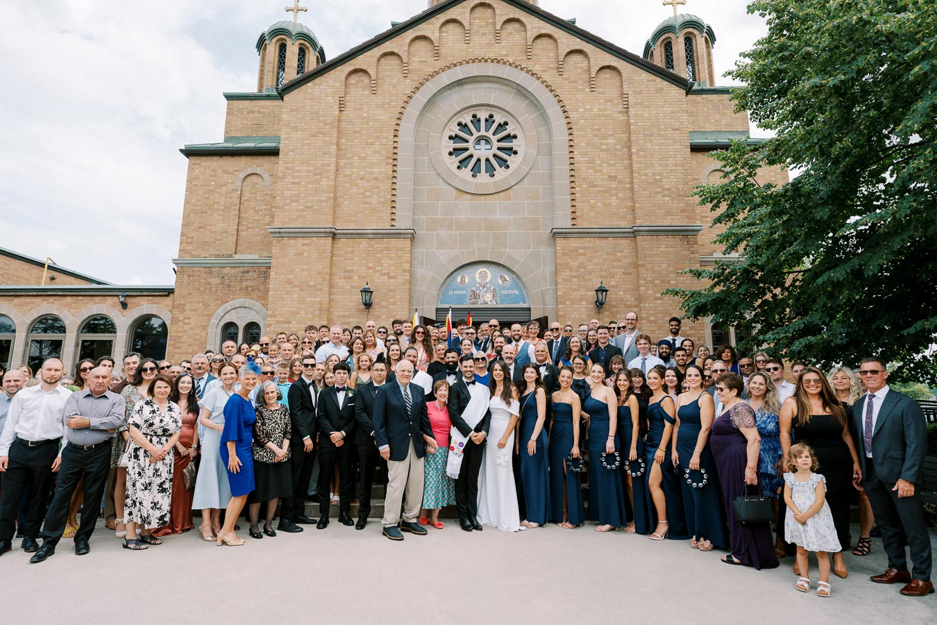 Group photo of wedding guests in front of a brick church building with large arched windows and a circular stained glass above the entrance. Guests, dressed in formal attire, gather around the bride and groom. Bridesmaids in navy dresses are positioned to the side, with trees partially visible.