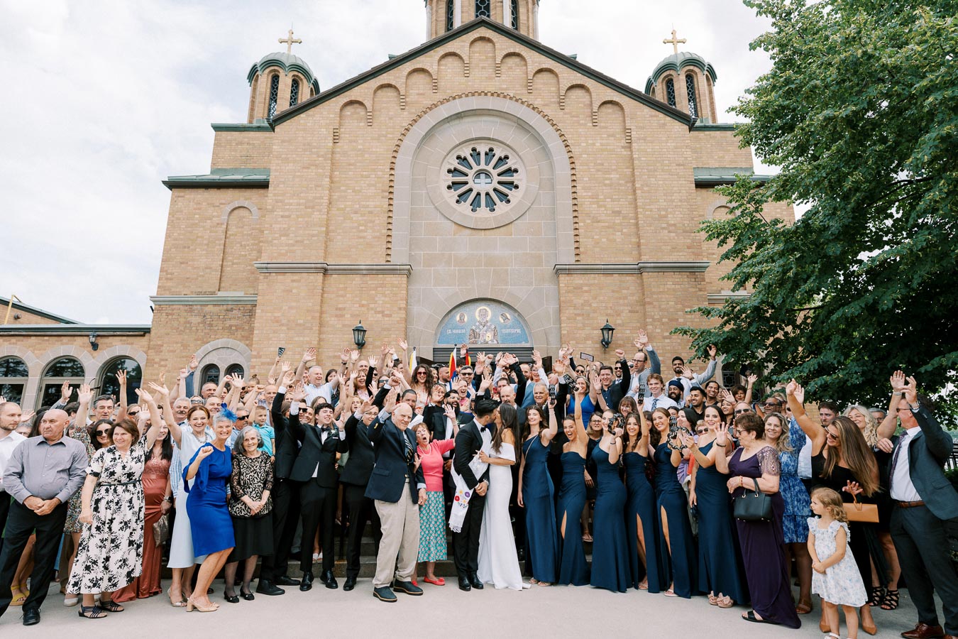 A large group of people celebrating a wedding in front of a historic church, with guests cheering and posing for a photo. The church's brick facade and ornate architecture are visible in the background, creating a festive and joyful atmosphere.