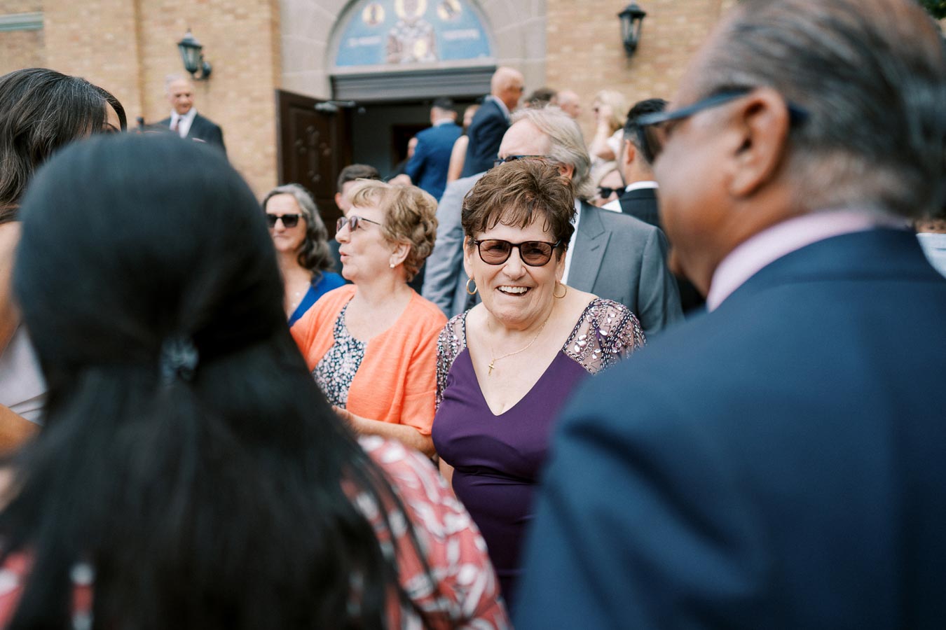 Group of people socializing outside a brick building during a formal event, with some attendees wearing sunglasses and elegant attire.