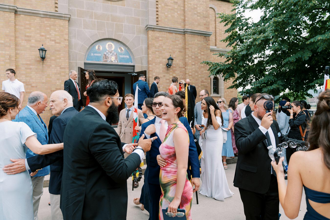 A group of people socializing outside a brick building, likely at a formal event. The crowd is dressed in suits and dresses, with some holding flags or capturing the moment with cameras. The atmosphere is lively and festive, indicative of a celebration or gathering, with architectural details of the building visible in the background.
