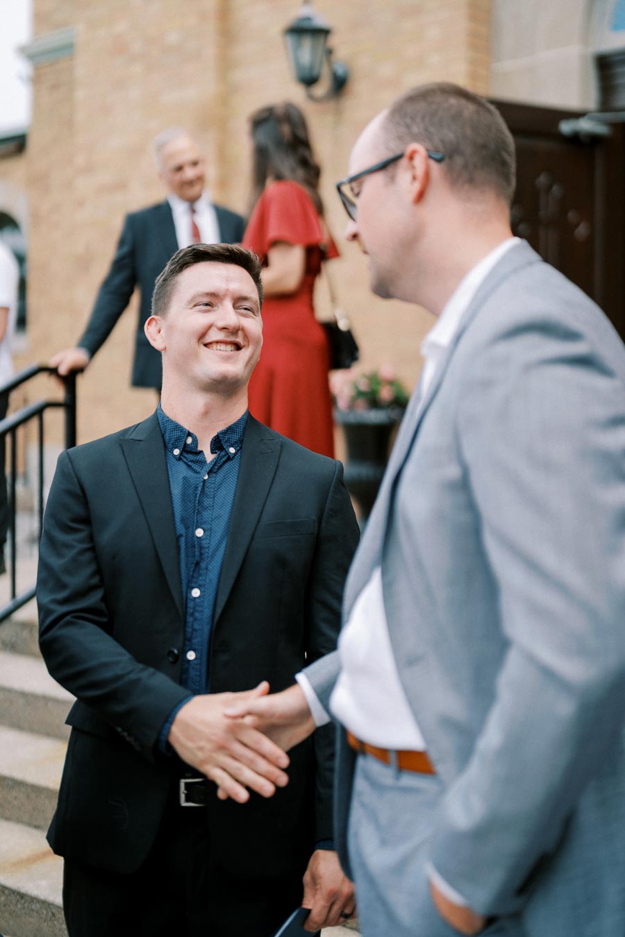 Two men in suits shaking hands and smiling outside a building, with others in the background.