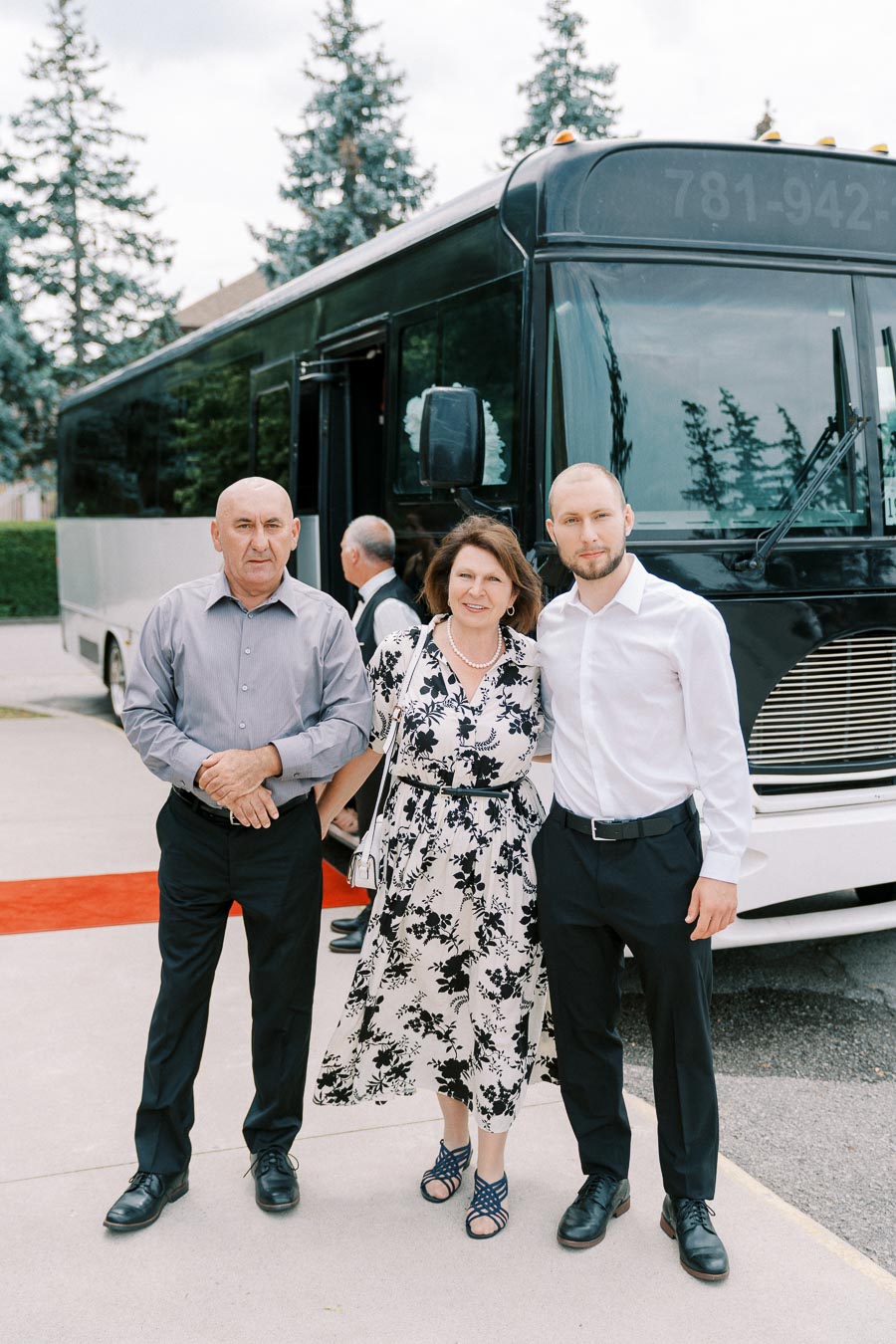 A group of three people smiling and posing in front of a black and white bus on a sunny day. Trees are visible in the background, and a red carpet leads to the bus entrance.
