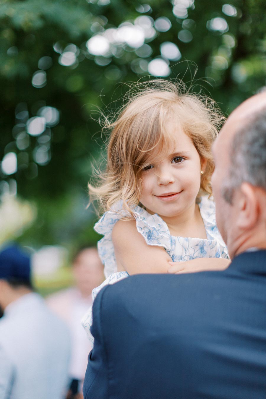 A young girl with wavy hair smiling while being held by an adult in a suit, surrounded by lush green foliage in an outdoor setting.