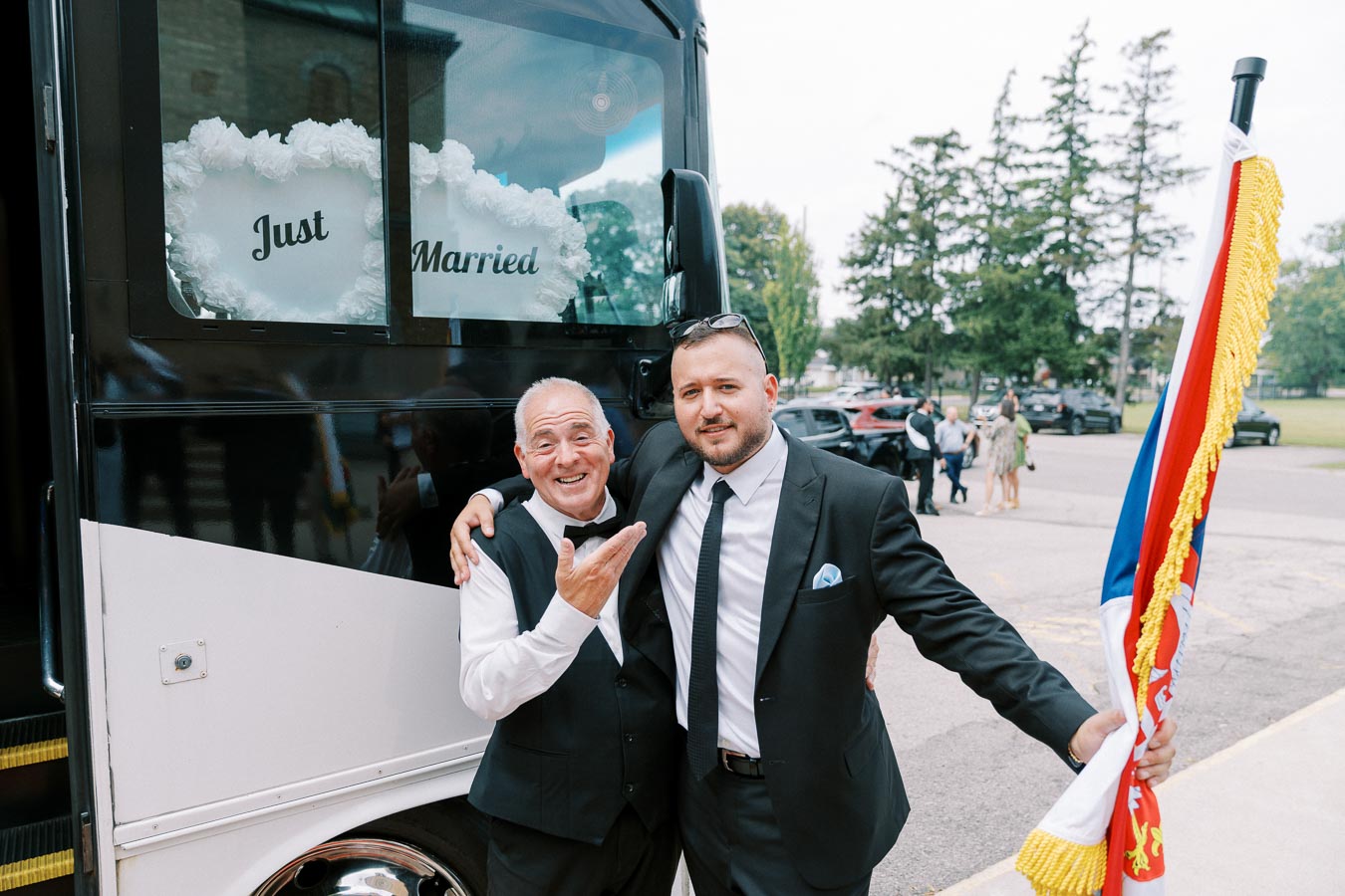 Two men in suits pose happily next to a Just Married decorated vehicle, one holding a colorful flag, outdoors with trees and people in the background.
