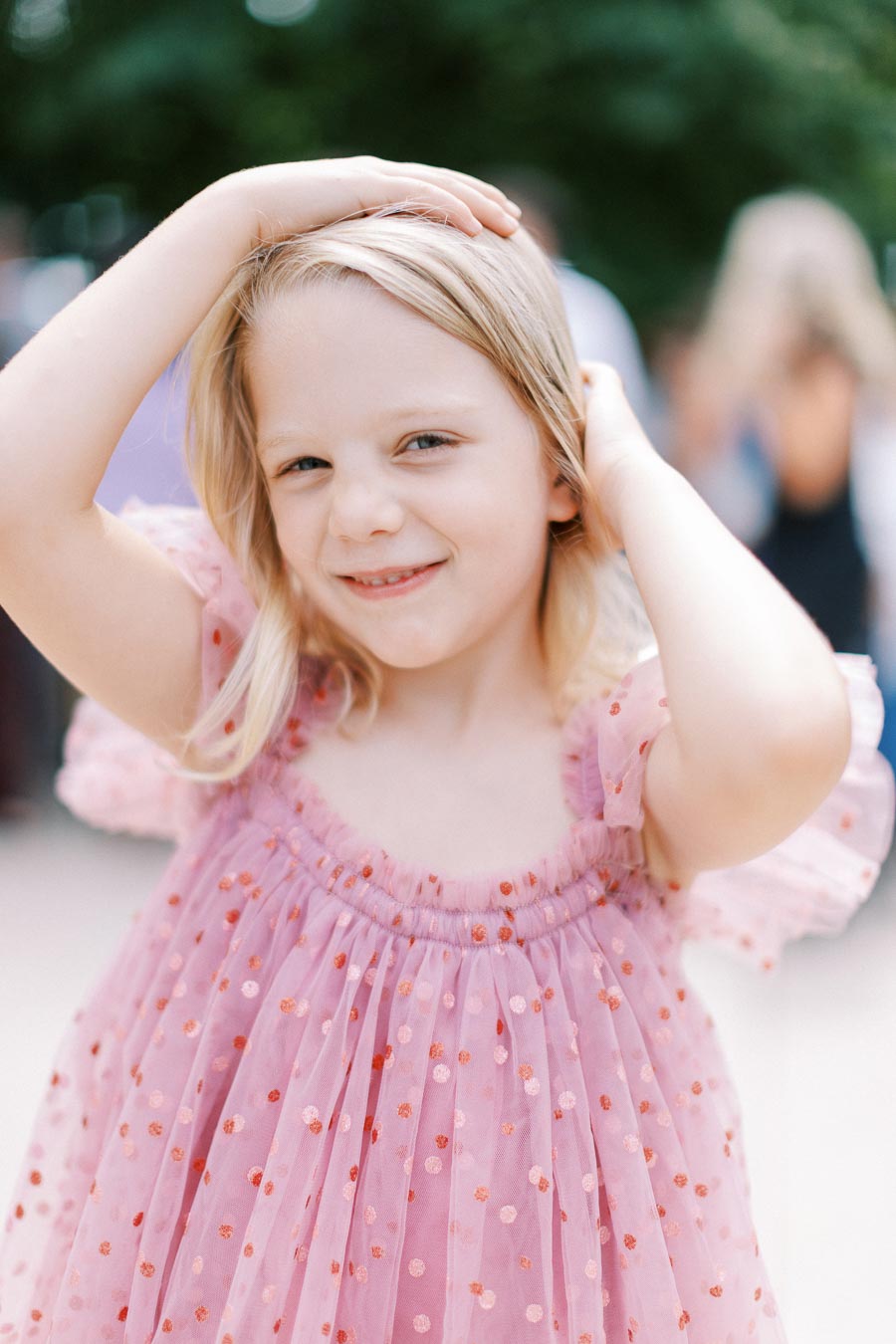 A young girl smiling and posing with hands on her head, wearing a pink polka dot dress in an outdoor setting.
