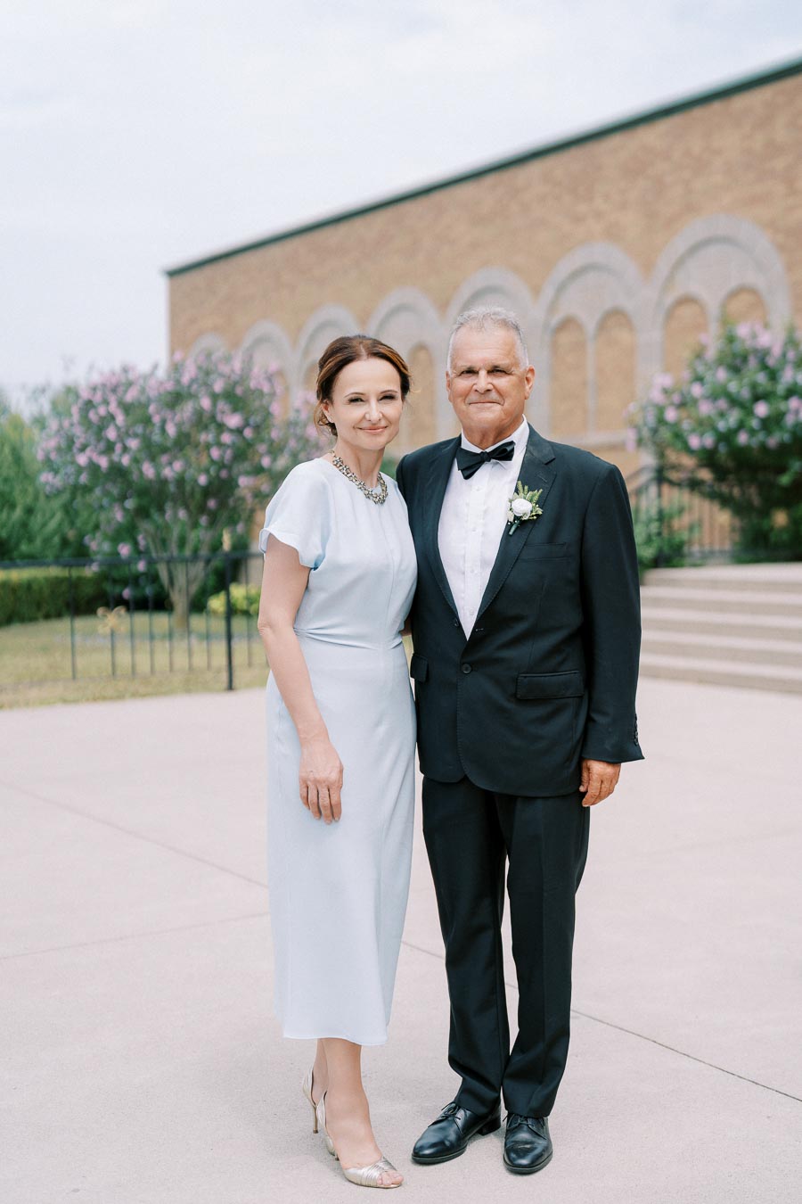 A well-dressed couple posing outdoors in formal attire, with a woman in a light blue dress and a man in a black suit and bow tie, in front of a beige building with arched windows and blooming trees.