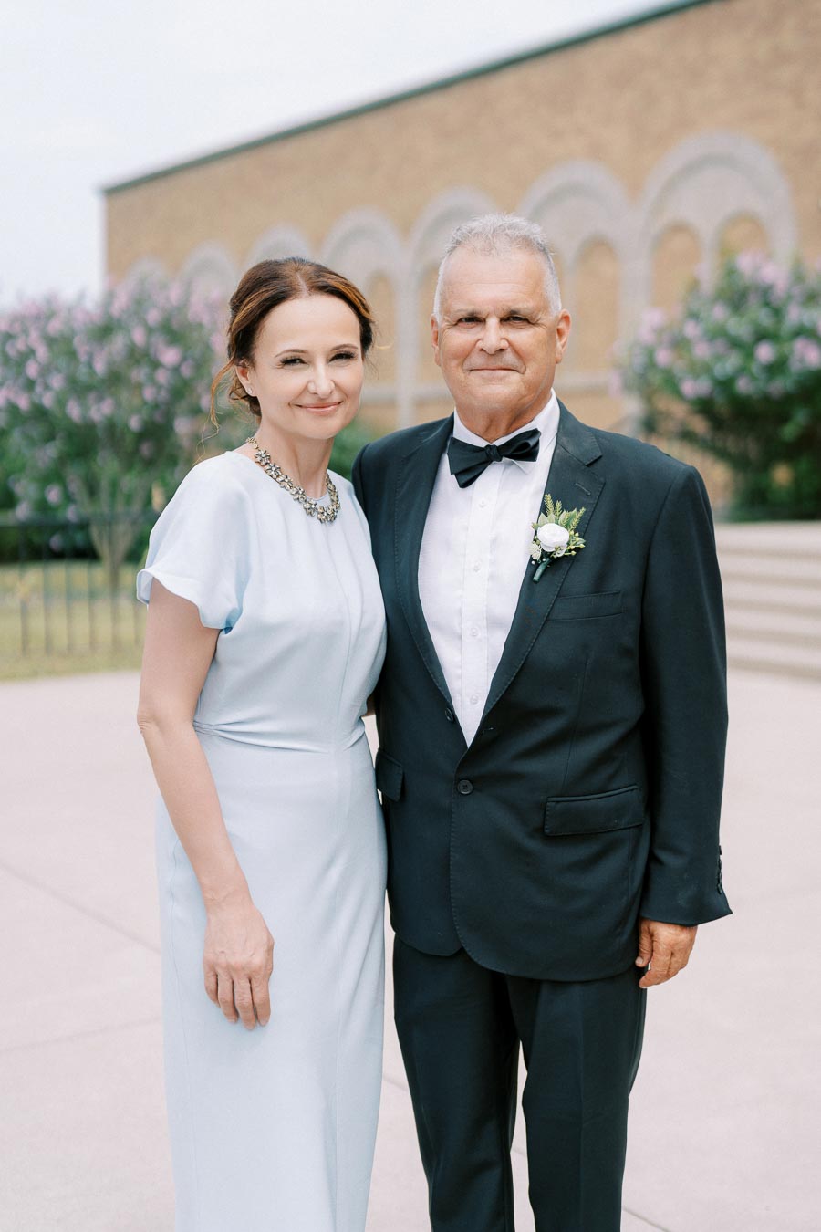 Mature couple in formal attire smiling outdoors against a backdrop of blooming trees and an elegant building.