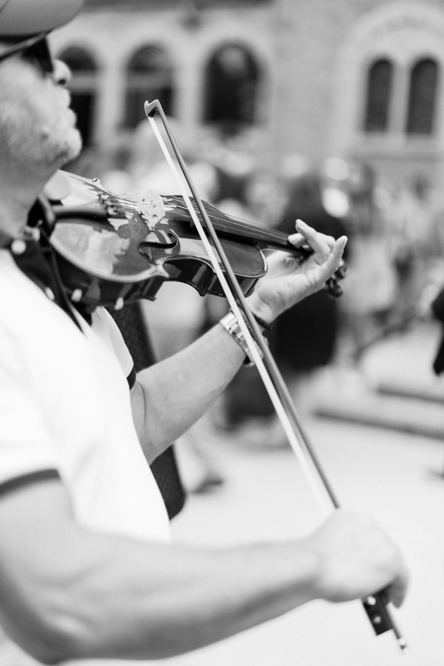 Black and white photo of a person playing a violin on a street, wearing a hat and sunglasses, with an arched building in the background.