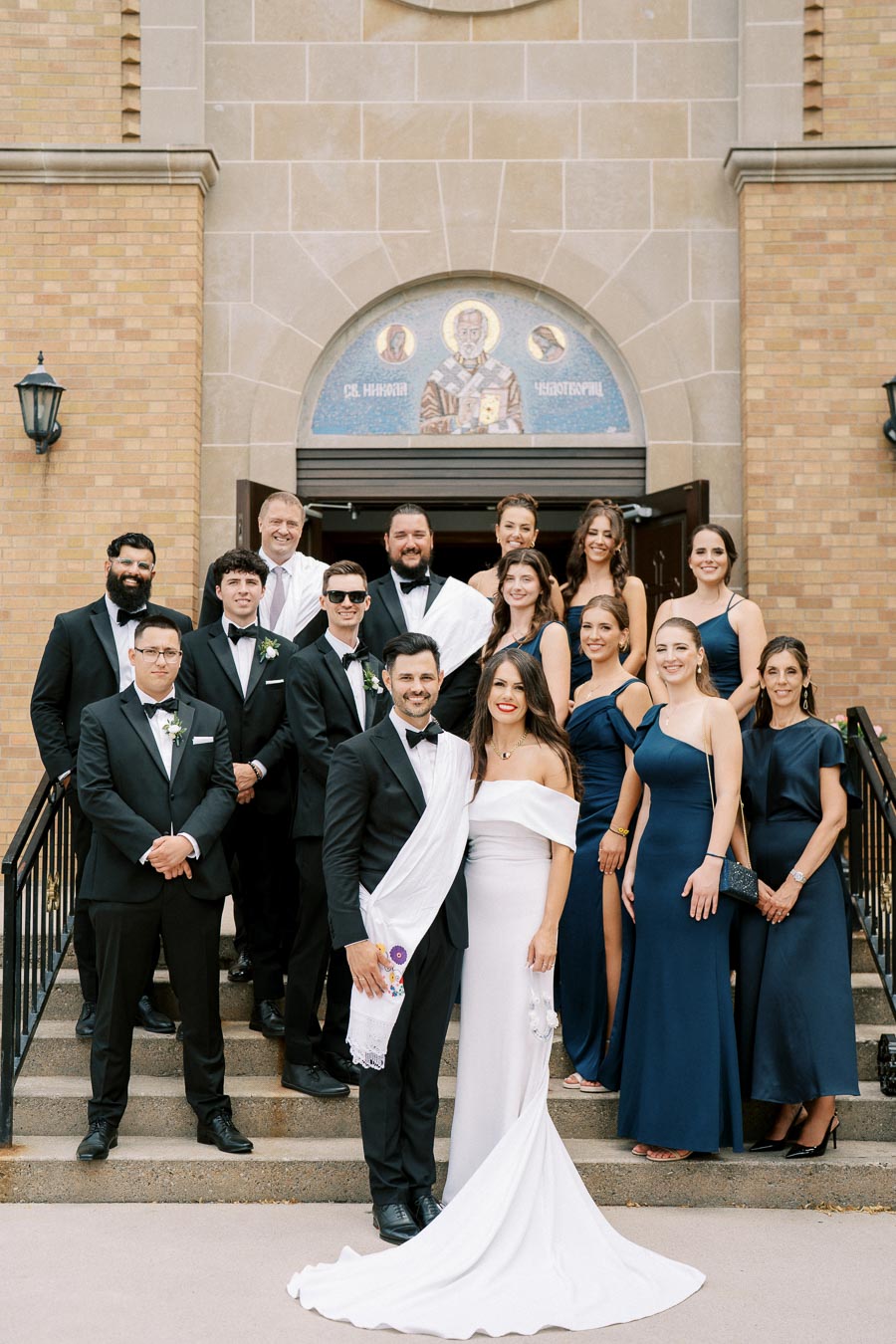 A bride and groom in elegant attire standing with a wedding party in formal dresses and suits on the steps of a church.