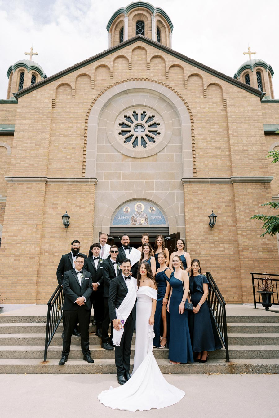 A wedding party poses on the steps of a historic church with intricate brickwork and round stained glass window, featuring a bride in a white gown and groom in a tuxedo, surrounded by bridesmaids and groomsmen in formal attire.