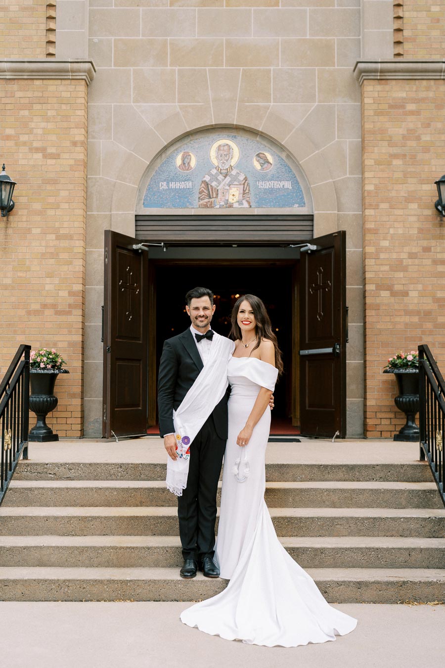 Wedding couple smiling outside a church entrance, groom in a black tuxedo and bride in a white gown, standing on steps with brick facade and religious iconography in background.