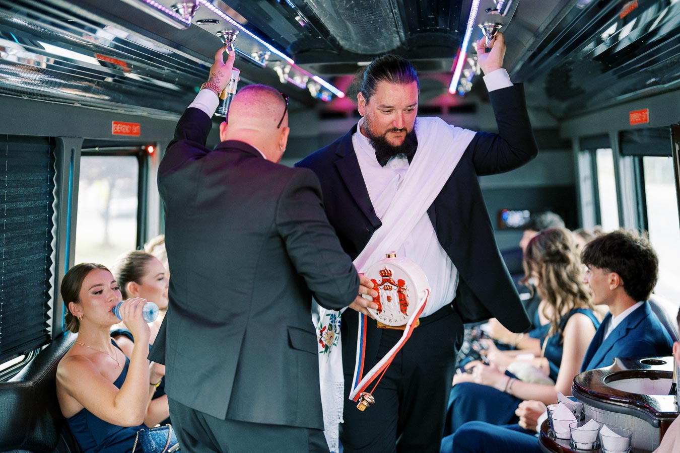 A group of people in formal attire enjoying a festive ride inside a party bus, with a focus on two men standing and holding drinks, and other passengers relaxing on the seats.