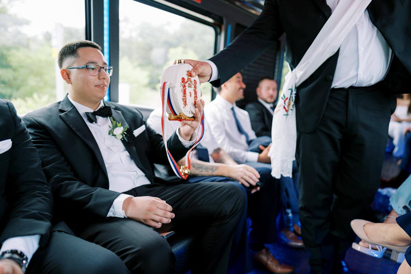 Groom in black tuxedo receives a traditional Serbian flask from a friend on a bus, surrounded by groomsmen in formal attire during a wedding celebration.