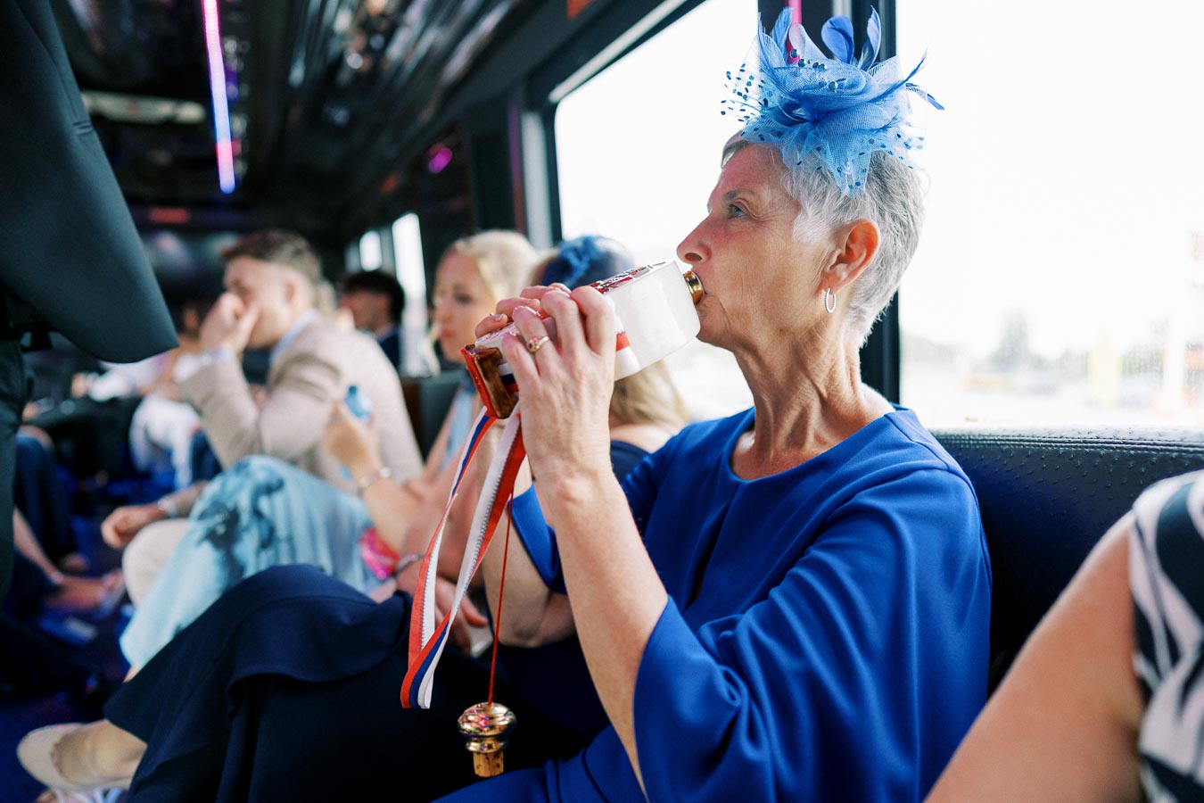Elderly woman in a blue dress and matching fascinator drinking from a flask on a bus, surrounded by formally dressed passengers.