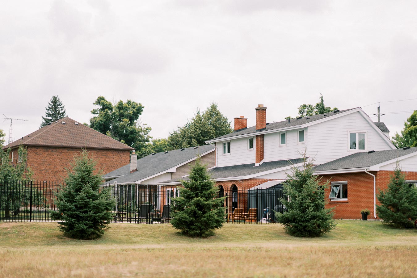 Suburban neighborhood with brick and white houses, fenced backyard, and evergreen trees under a cloudy sky.