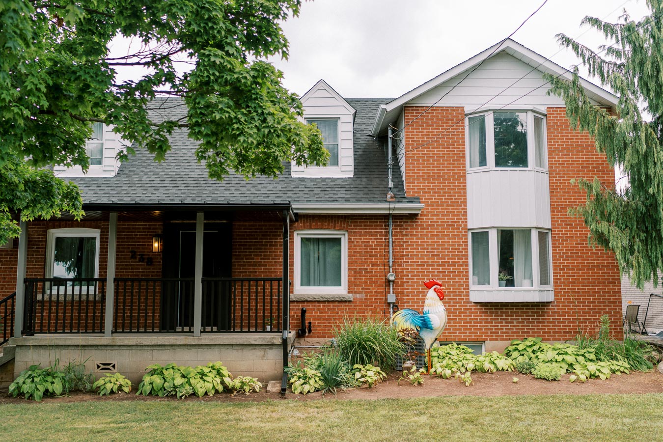 Brick house with a large front porch, lush greenery, and a colorful rooster statue in the garden, under a cloudy sky.