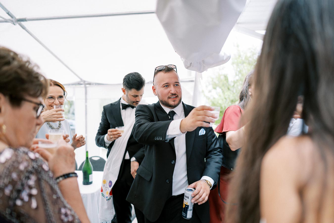 A group of elegantly dressed people socializing and toasting with drinks at a formal event under a white tent.