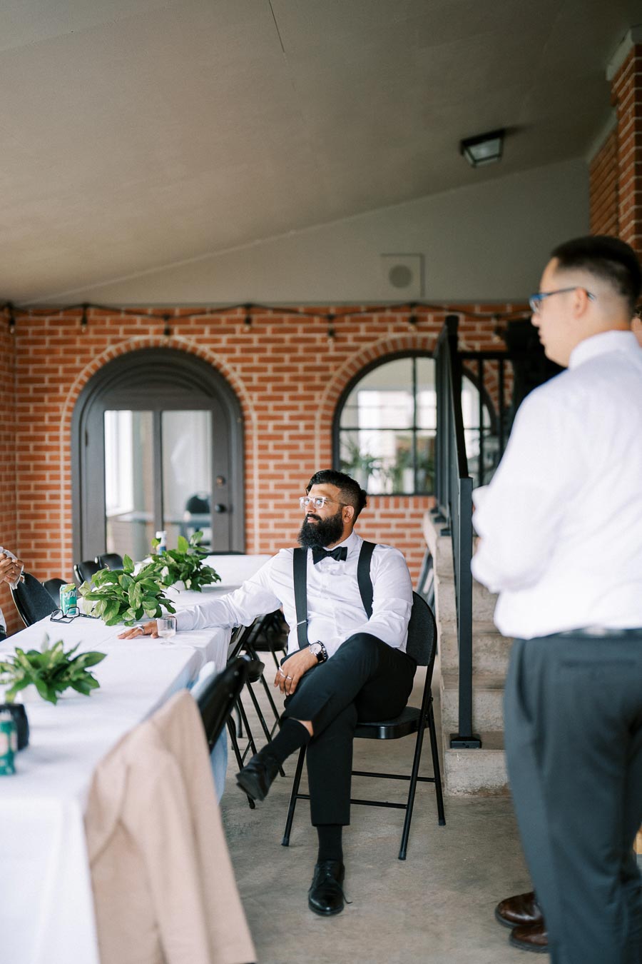 Man in formal attire with suspenders and bow tie sitting at a white table with greenery in a modern brick interior.