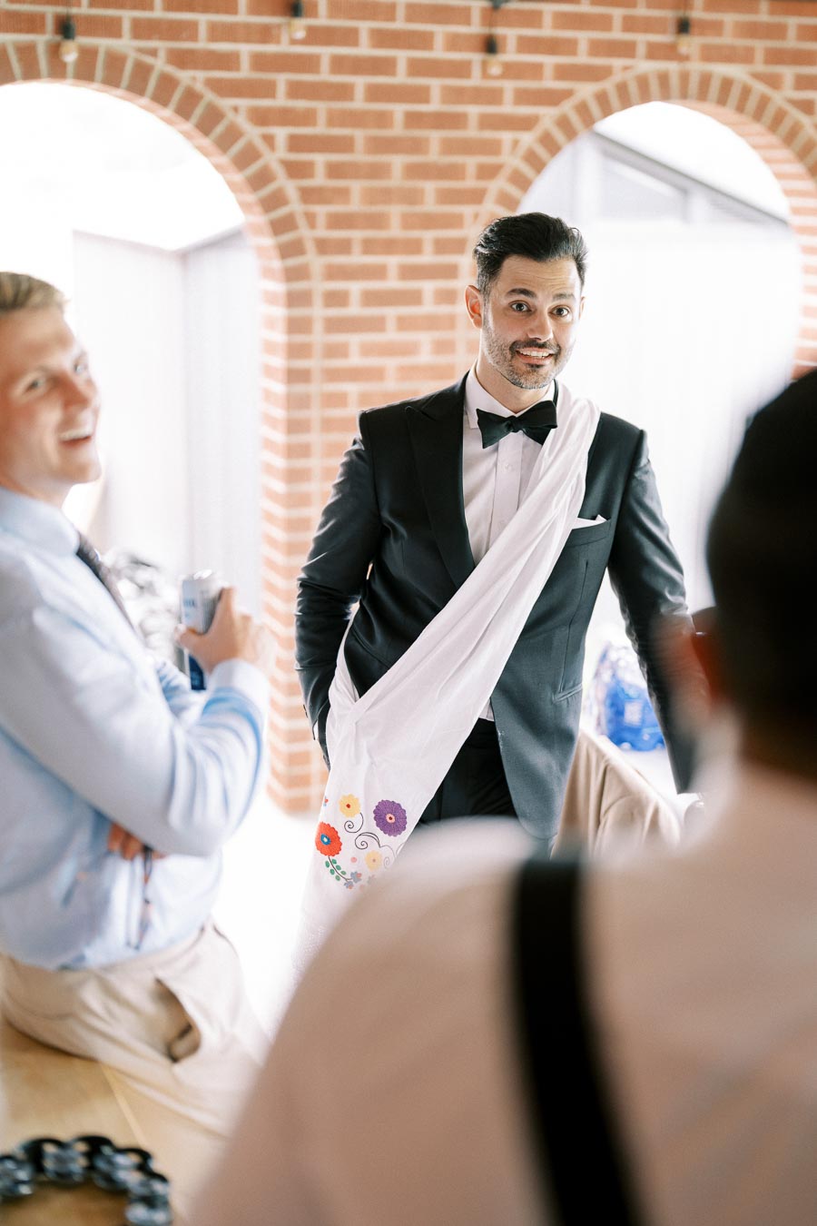 A man in a black tuxedo and bow tie stands smiling indoors, with a white floral embroidered cloth draped over his shoulder.