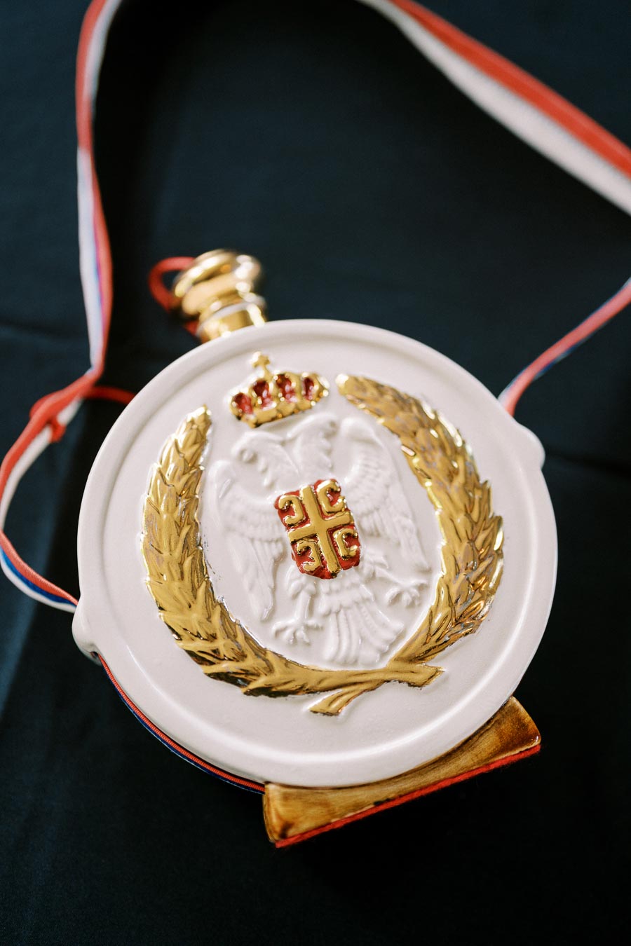 A decorative white flask with a gold emblem and wreath, featuring a crown and red details, on a dark surface with a tricolored strap.