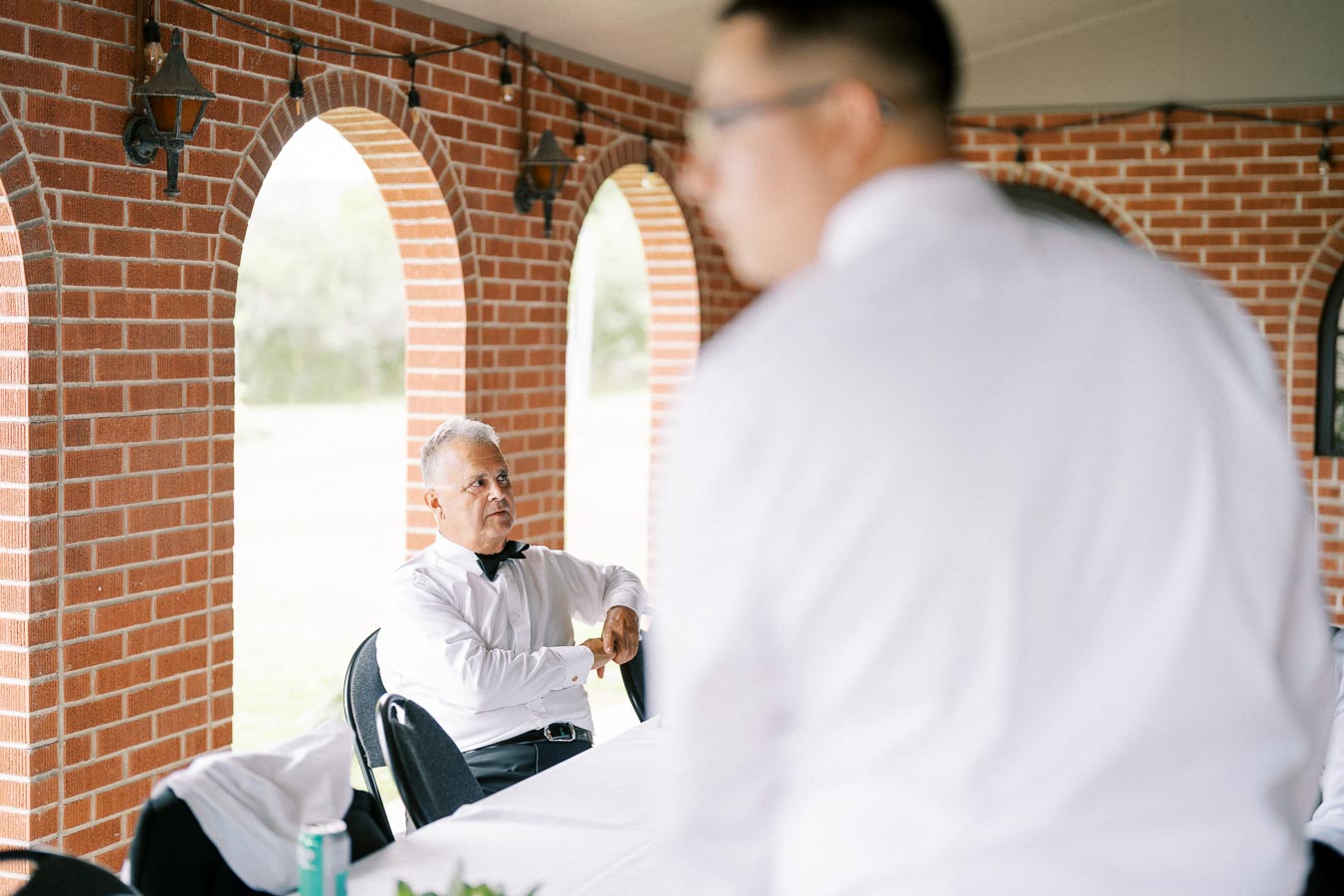 Senior man in a tuxedo sitting at a white-clothed table, under an arched brick pavilion, with another person standing nearby in focus.