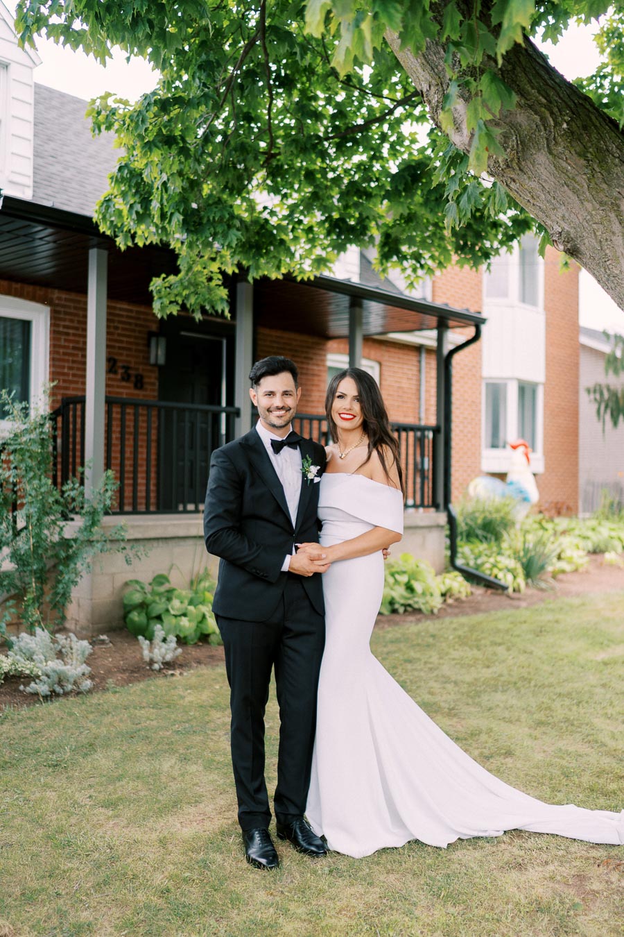 A couple in formal attire posing outdoors in front of a brick house with a lush garden, featuring a man in a black suit and a woman in an off-shoulder white gown.