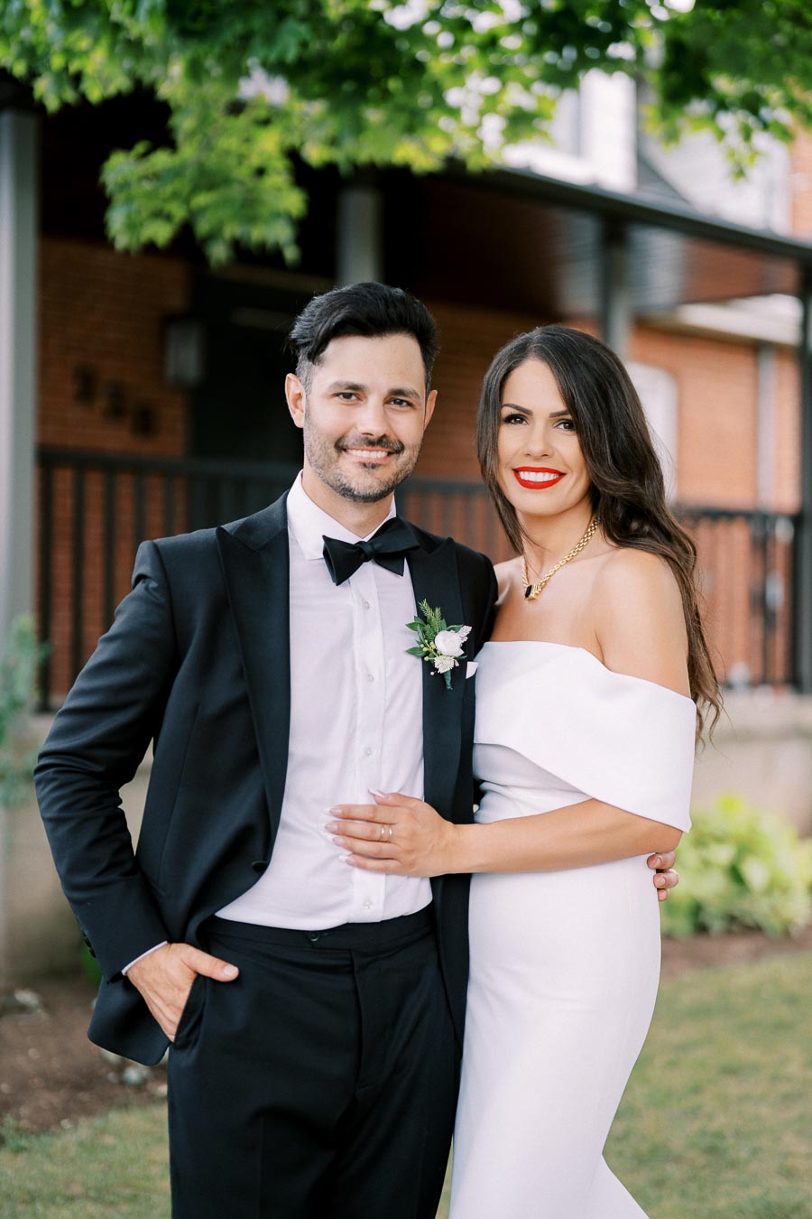 Smiling bride and groom in elegant wedding attire posing outdoors with lush greenery in the background.