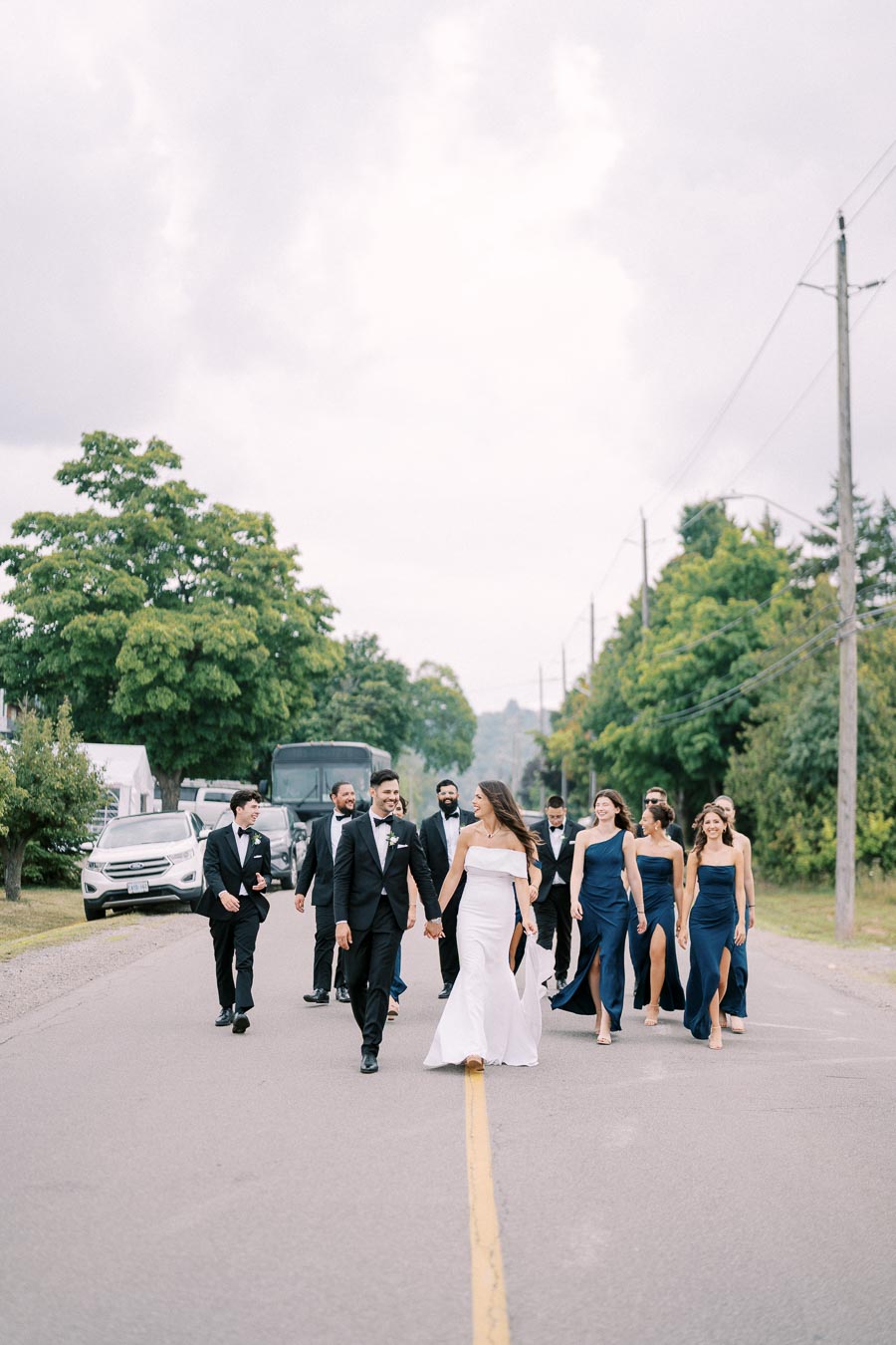 Bridal party walking along a tree-lined road, with the bride in a white dress and bridesmaids in navy gowns, set against a cloudy sky.