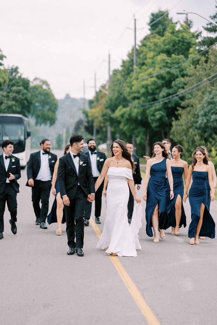 A wedding party walking down a paved road, featuring a bride in a white gown and groom in a black suit, surrounded by bridesmaids in blue dresses and groomsmen in black suits on a tree-lined street.