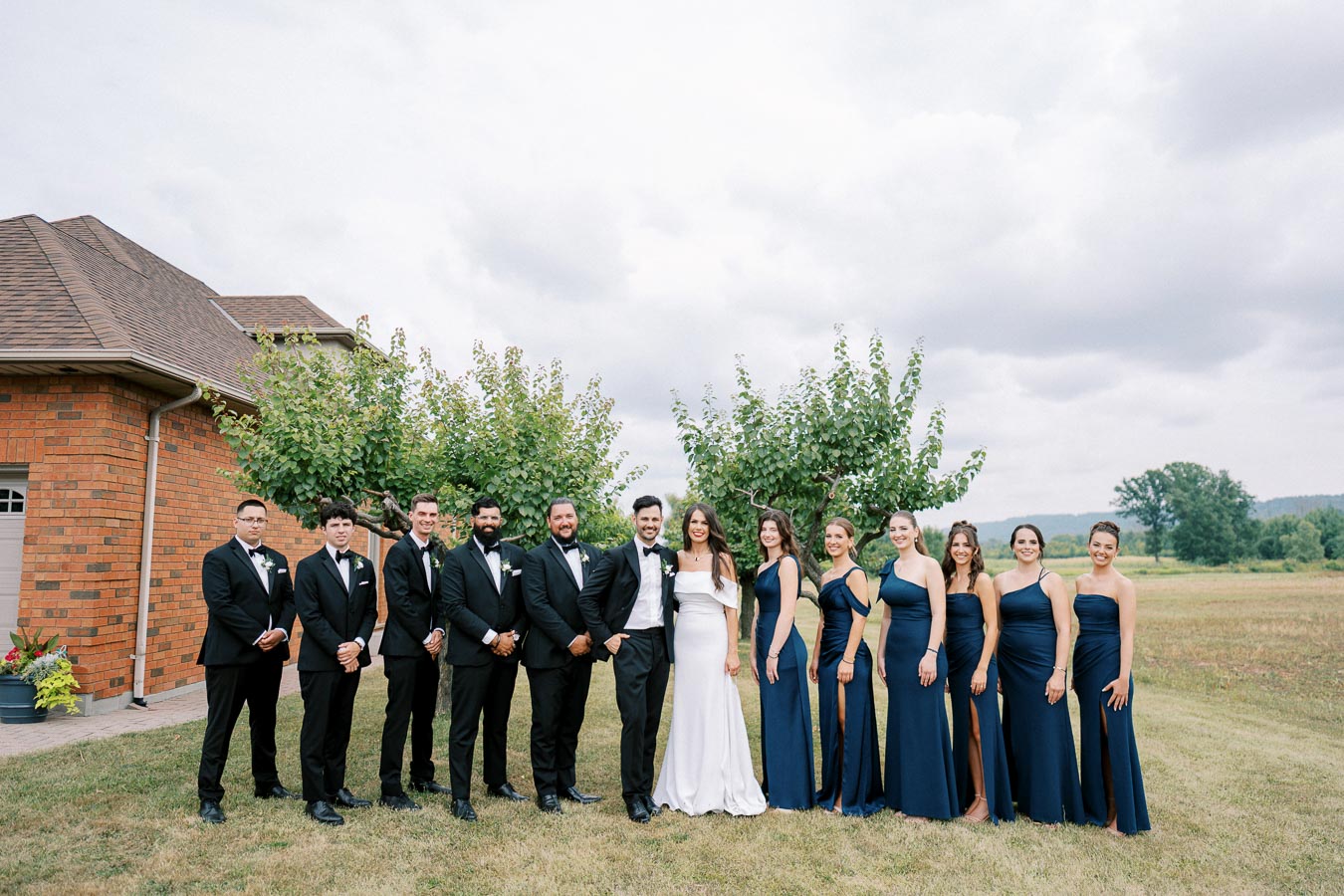 A wedding party posing outdoors, featuring a bride in a white gown and groom in a tuxedo with bridesmaids in navy blue dresses and groomsmen in black suits, standing in front of a brick house and greenery under a cloudy sky.