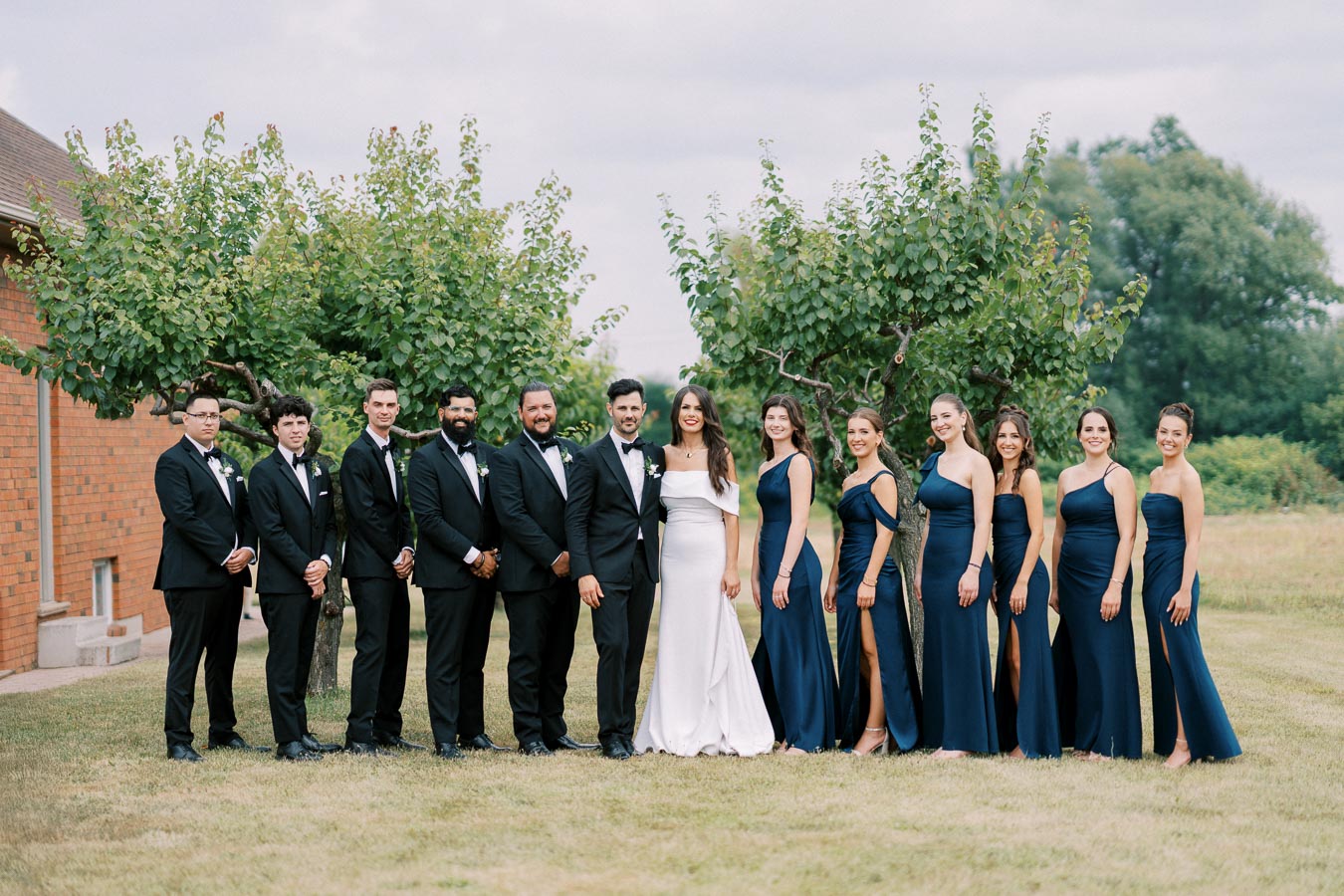 Wedding party posing outdoors with bridesmaids in navy blue dresses and groomsmen in black suits, standing on a grassy lawn with trees and a brick building in the background.
