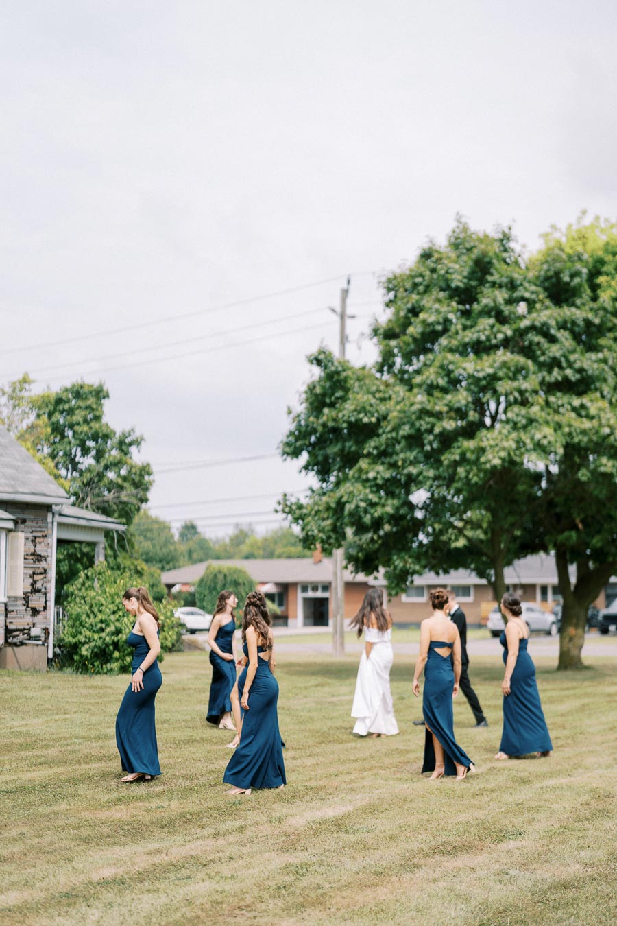 A group of bridesmaids in navy blue dresses and a bride in a white gown walk across a grassy lawn, surrounded by trees and houses under a cloudy sky.