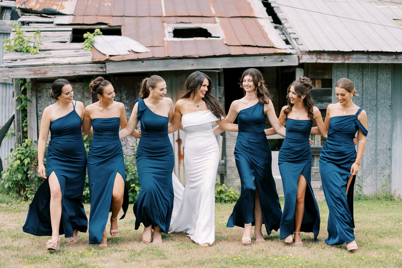 Bridal party in matching navy blue dresses and white bridal gown walking together outside in front of rustic barn, smiling and linking arms.