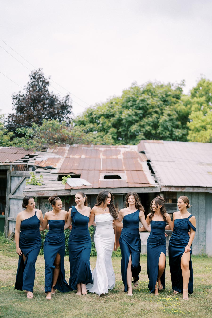 Bride in white dress walking with bridesmaids in matching blue dresses, posing outdoors in front of rustic barn.