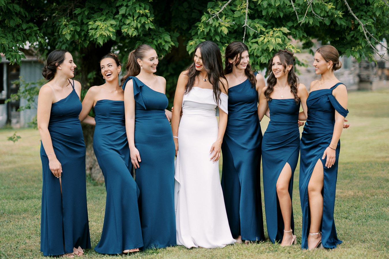 Bridal party posing outdoors with bridesmaids in matching blue dresses and the bride in a white gown, under a leafy tree background.