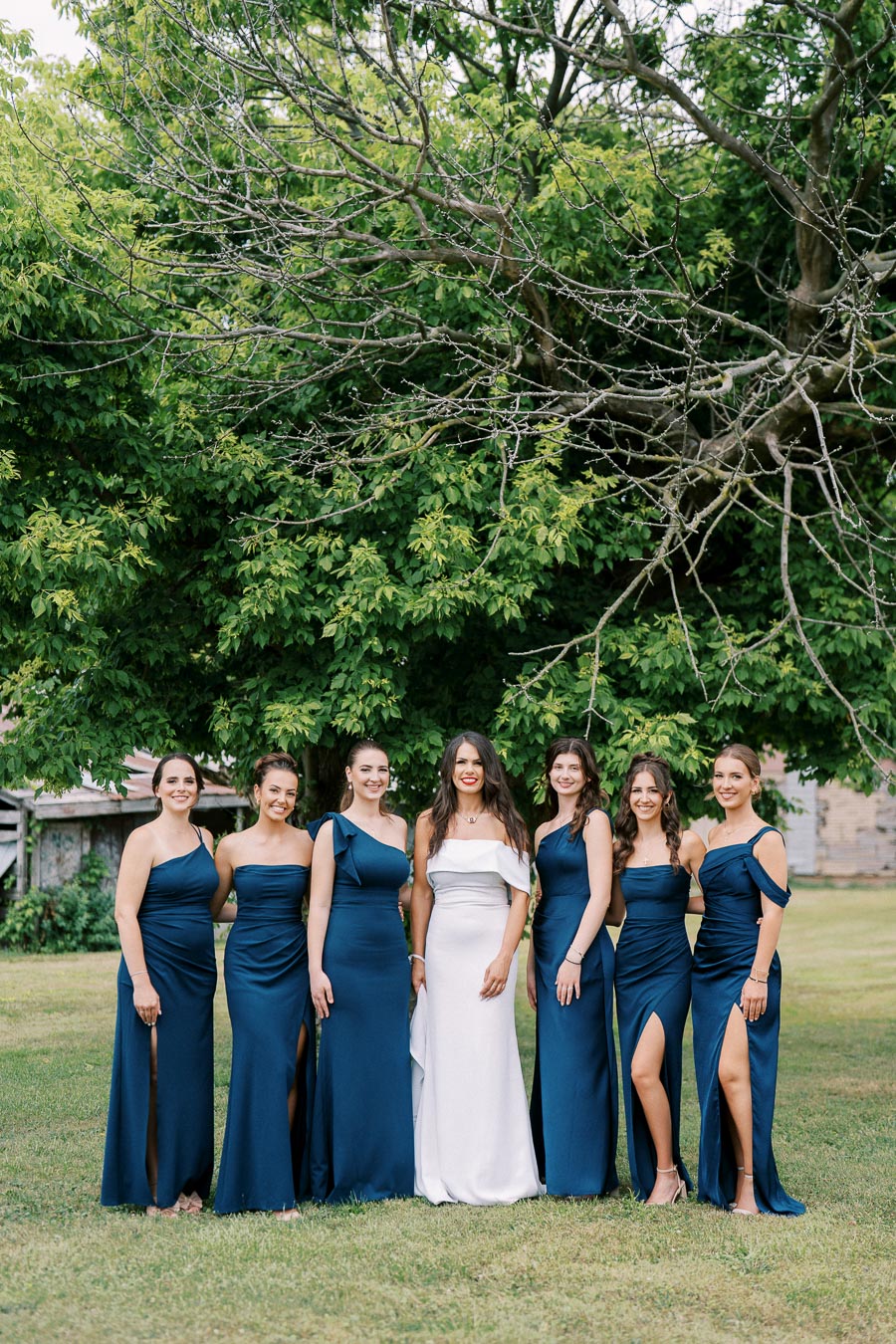 Bridal party posing outdoors against a backdrop of lush greenery, with bridesmaids in elegant navy blue dresses and the bride in a white gown, showcasing formal wedding attire.