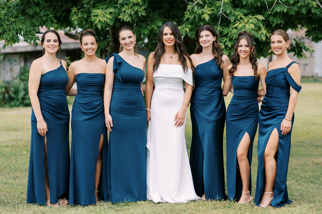 A bride in a white dress stands in the center, surrounded by six bridesmaids wearing matching teal gowns, posing outdoors in front of lush green trees.