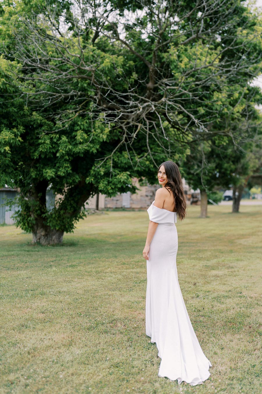 A woman in an elegant off-the-shoulder white dress poses outdoors, surrounded by lush green trees and grass, capturing a serene and picturesque moment.