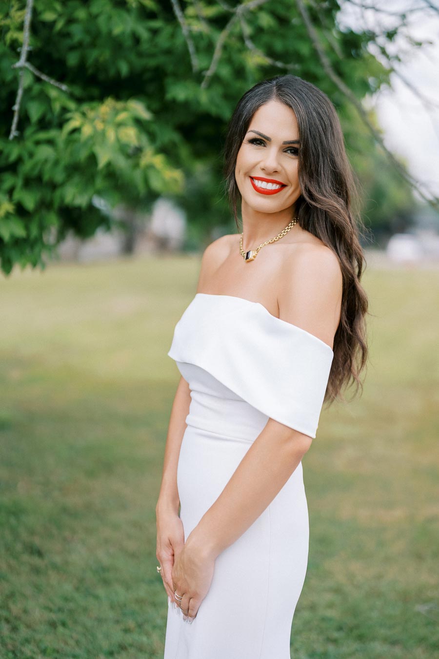 A woman in an elegant off-shoulder white dress smiles in an outdoor setting with lush green trees in the background.