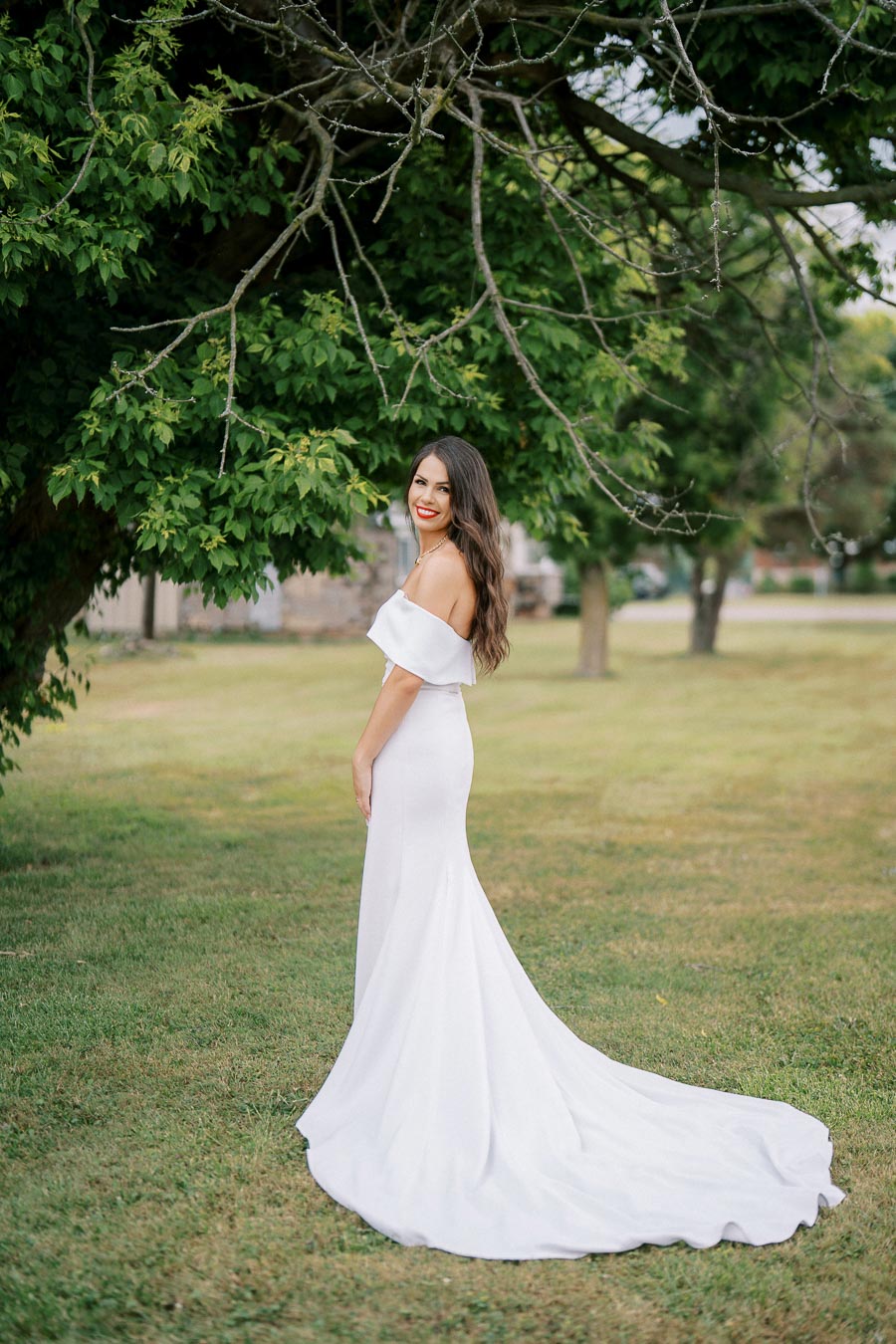 A woman in an elegant white off-shoulder dress stands on green grass, smiling under the branches of a lush tree. The scene captures a serene outdoor moment perfect for a bridal photoshoot.
